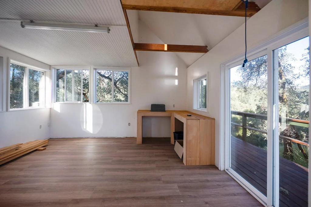 Empty room with large windows, a sliding glass door leading to a balcony, wood flooring, a built-in wooden desk, and a ceiling with exposed beams.