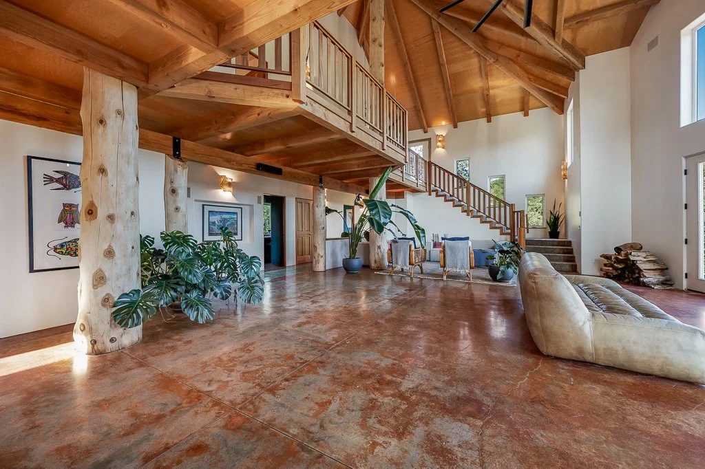 Spacious living room with wooden beams and high ceilings, featuring a beige sofa, potted plants, artwork on the walls, and a staircase leading to an upper level.