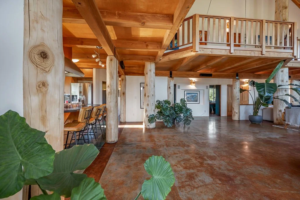 Spacious open-concept interior with wooden beams, wood-paneled ceiling, and polished concrete floors, featuring a bar area with chairs, indoor plants, and a second-floor balcony.