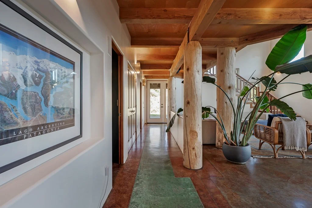 Interior of a modern home with wooden beams and columns, a large potted plant, a framed map of Argentina Patagonia on the wall, a green rug on the floor, and a cozy sitting area with wicker chairs and a blanket.
