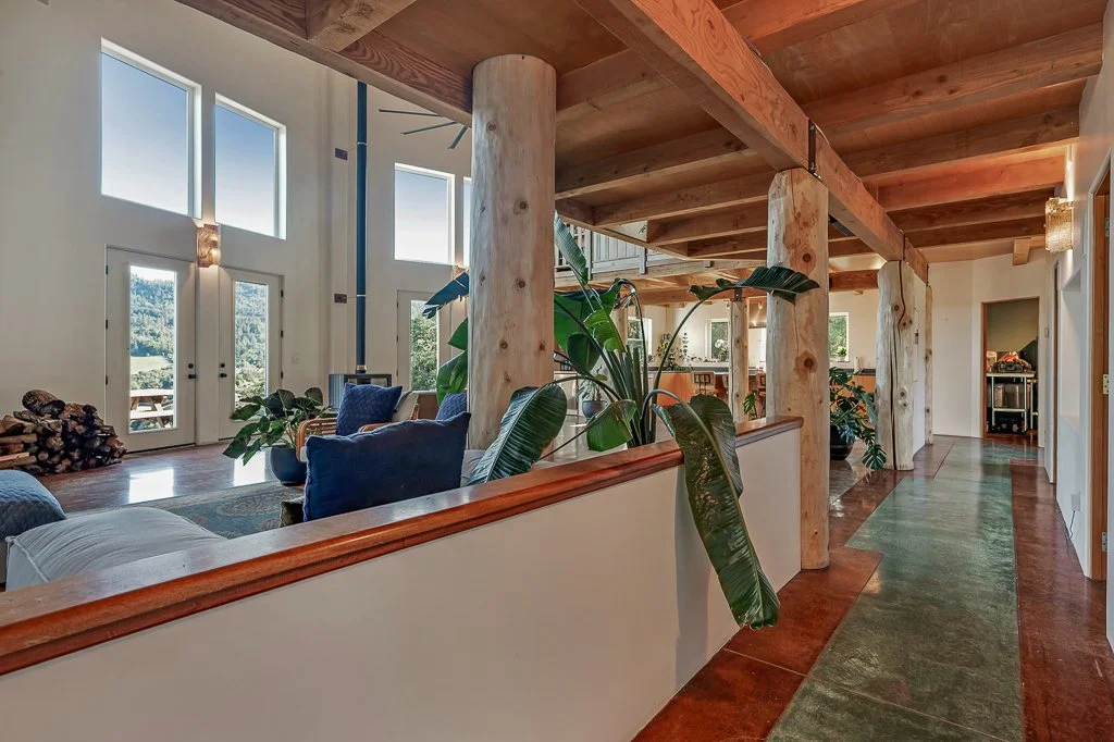 Open living room with large windows, wooden beams, and modern decor, featuring a cozy seating area, plants, and a view of trees outside.