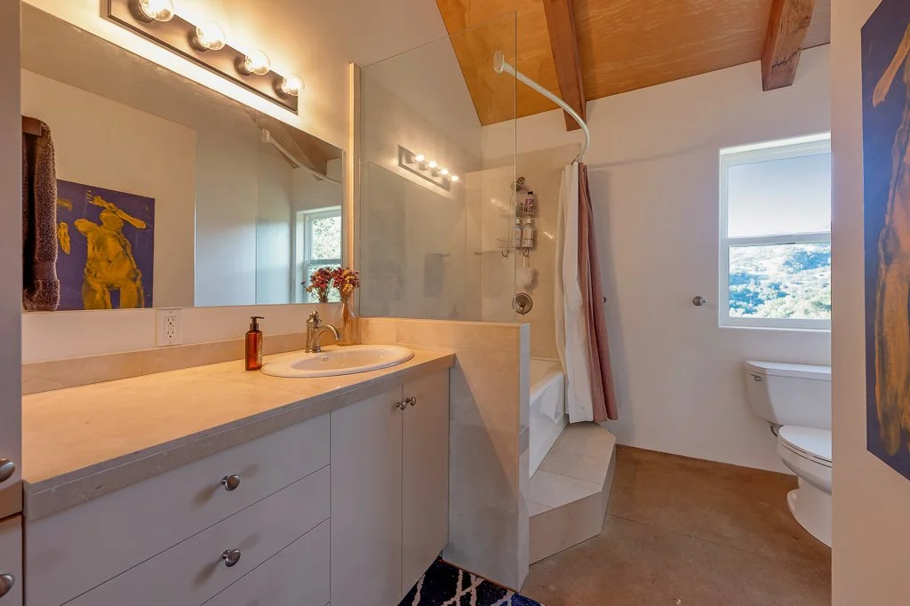 Bathroom with a large mirror above a beige countertop with a sink, small flower vase, and soap dispenser. To the right, a shower area with a curved shower rod and curtain, a window showing a scenic view, and a toilet. The ceiling is wooden with expos