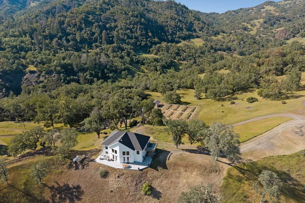 An aerial view of a white house with a dark roof in a lush, green hilly landscape with trees and open grassy areas