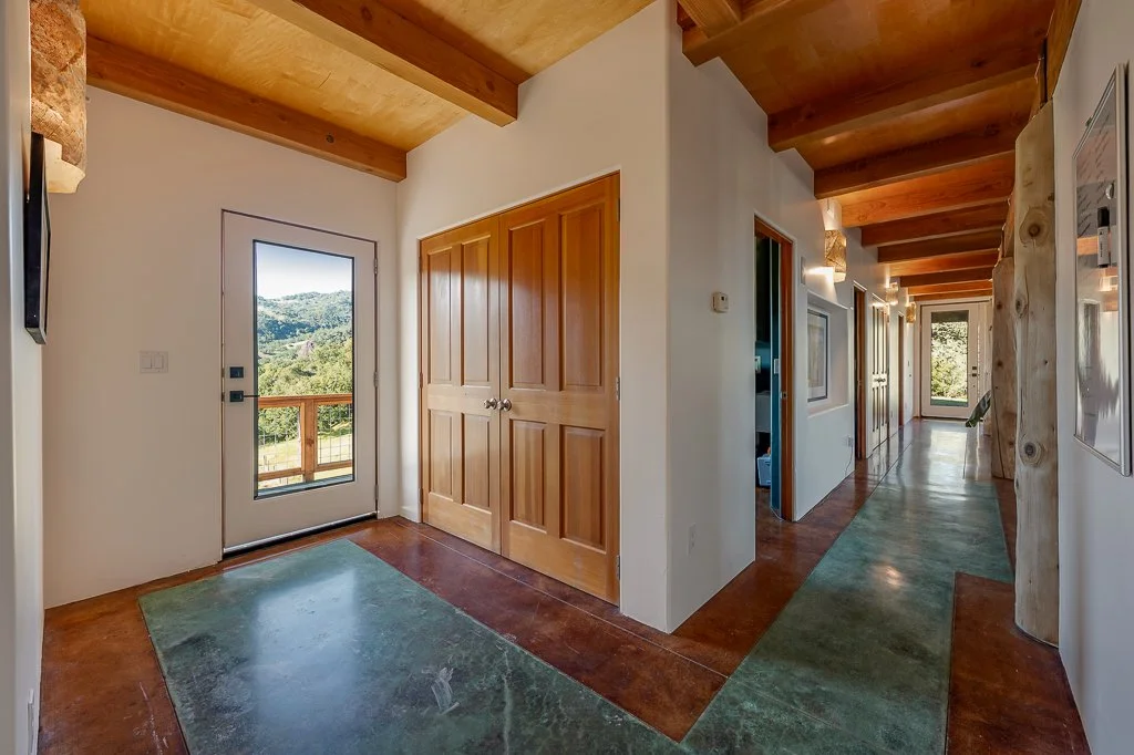 Interior view of a hallway in a house with a wooden ceiling and door frames, white walls, and a concrete floor with green patches, leading to a door with a view of a scenic landscape outside.