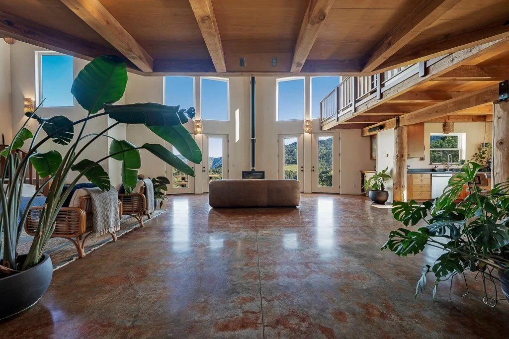 Spacious living room with high ceilings and large windows showing views of greenery and mountains, featuring wooden beams, a brown ottoman, wicker chairs, potted plants, and a kitchen area in the background.