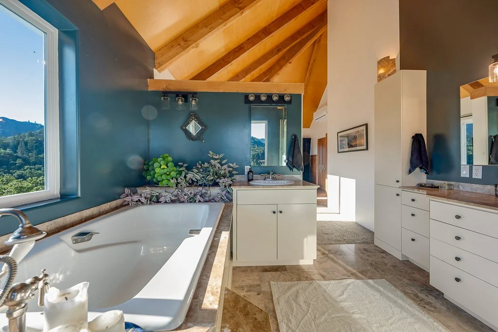 Bathroom with a large corner tub, white cabinetry, blue and white walls, window overlooking a mountain landscape, wooden ceiling beams, and a mirror above the sink.