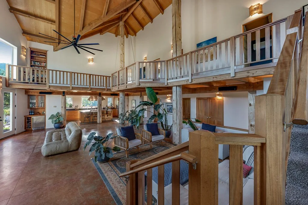 Spacious living room with a two-story ceiling featuring exposed wooden beams and a modern black ceiling fan. The area includes a cozy cream-colored sofa, multiple blue and wooden chairs, and several plants. The room opens to a kitchen with a large wi