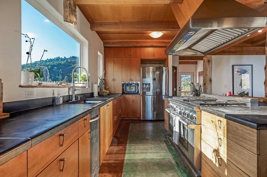 Kitchen with wooden cabinets, black countertop, stainless steel appliances, large window with mountain view, and wooden ceiling beams.