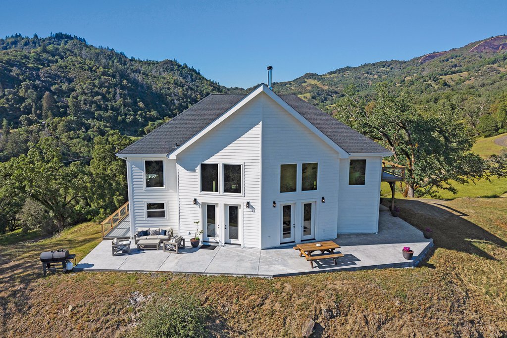 White two-story house with large windows, outdoor patio with seating, picnic table, grill, and potted plants, set in a green hilly landscape under a clear blue sky.