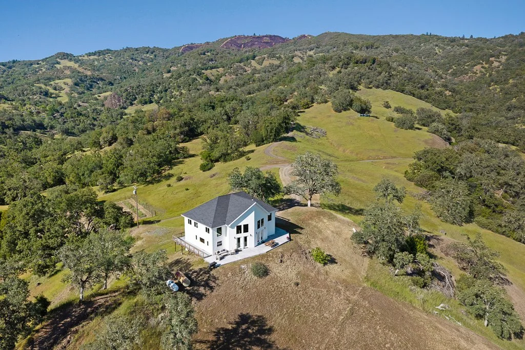 A white two-story house with a deck, situated on a hillside surrounded by trees and rolling green hills under a clear blue sky.