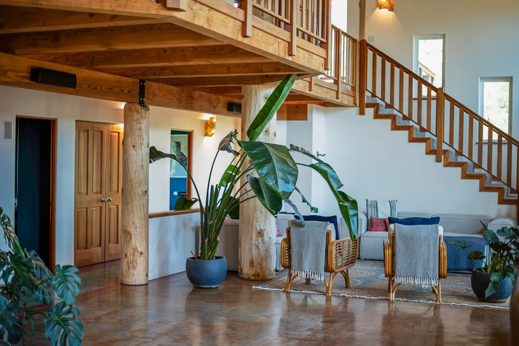 Interior of a spacious living room with wooden beams, large potted plants, and furniture including a beige sectional sofa and two rattan chairs with cloth covering, illuminated by natural light from windows.