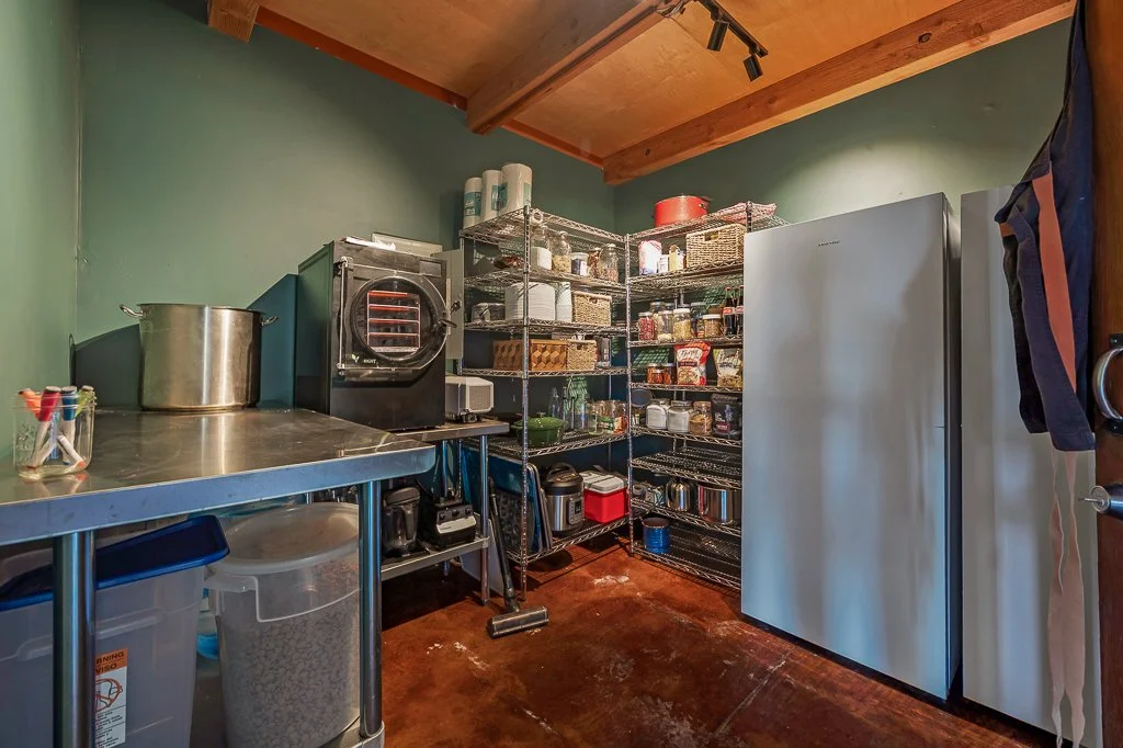 A pantry with metal shelving filled with canned goods, jars, and kitchen supplies, a large refrigerator, and a stainless steel counter with a pot and appliances.