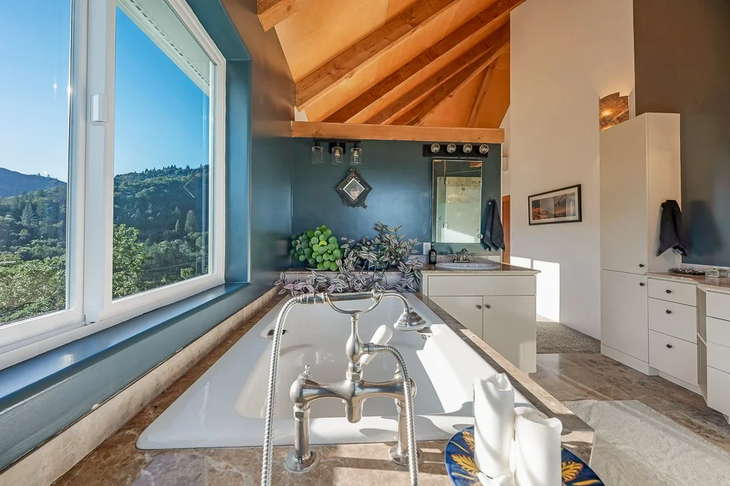Interior view of a kitchen with a large window showing a scenic mountain landscape, a bathtub with vintage faucet fixtures, and white cabinetry with a mirror and framed artwork