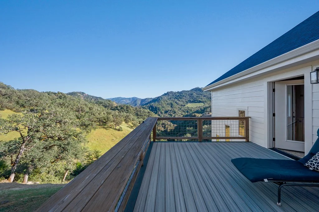 A wooden balcony with a lounge chair overlooking green hills and mountains under a clear blue sky.