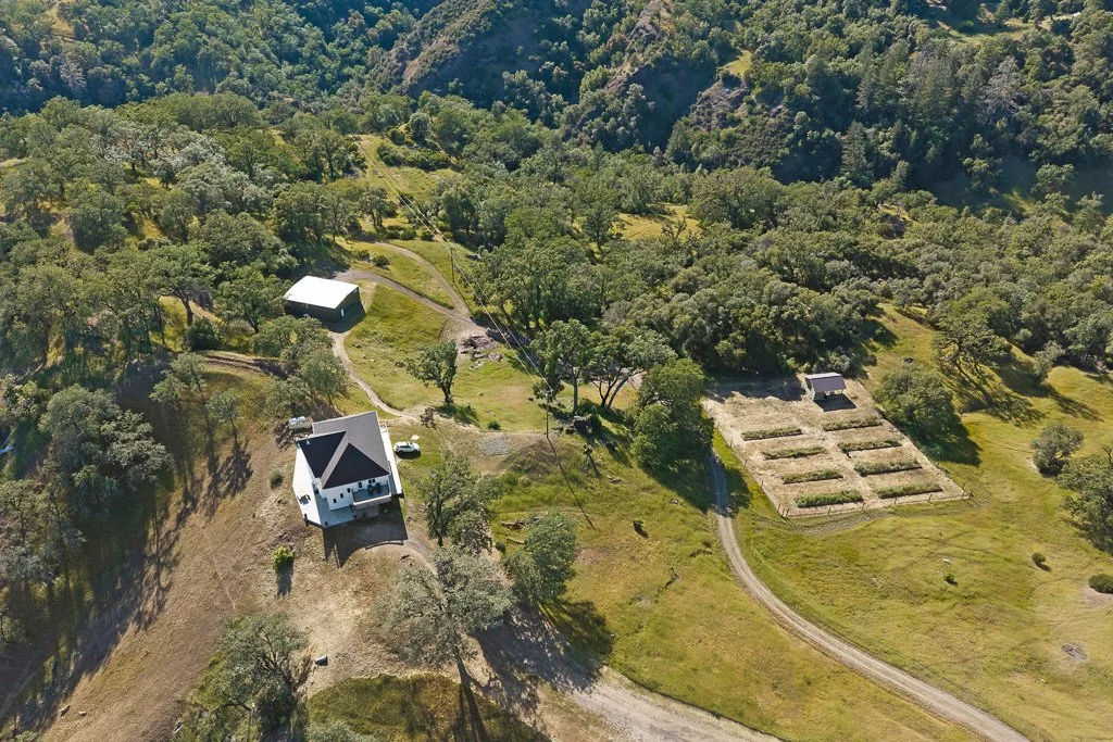 Aerial view of a rural property surrounded by dense trees and greenery, showing a white house, a dirt road, a large metal shed, and a rectangular garden layout.