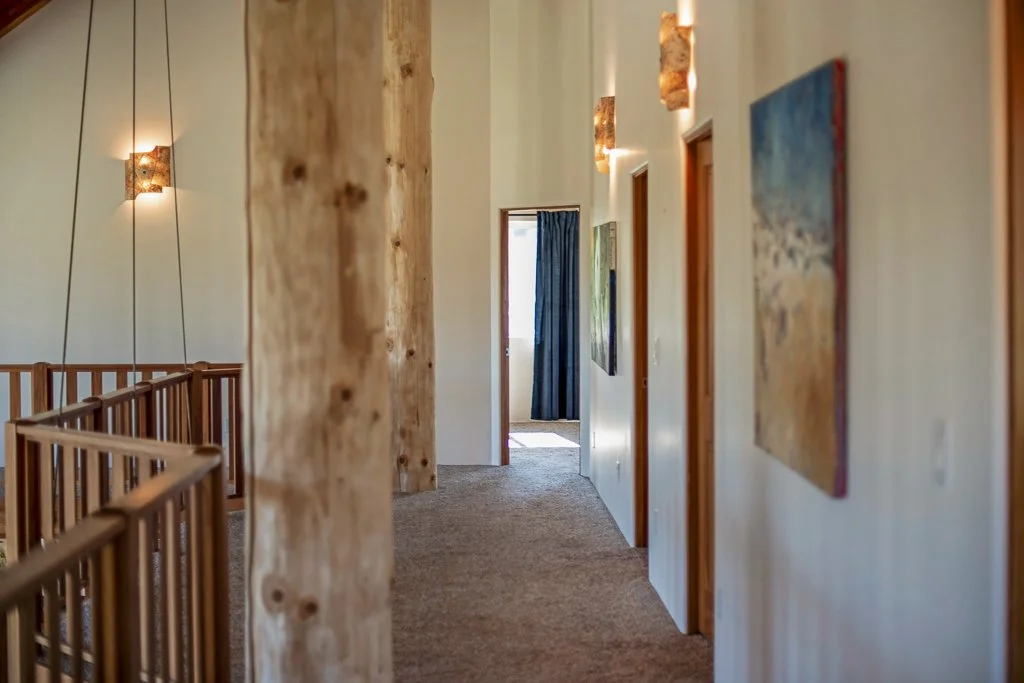A hallway in a house or lodge with wooden posts, wall-mounted lamps, and artwork, leading to a partially open door with a blue curtain.