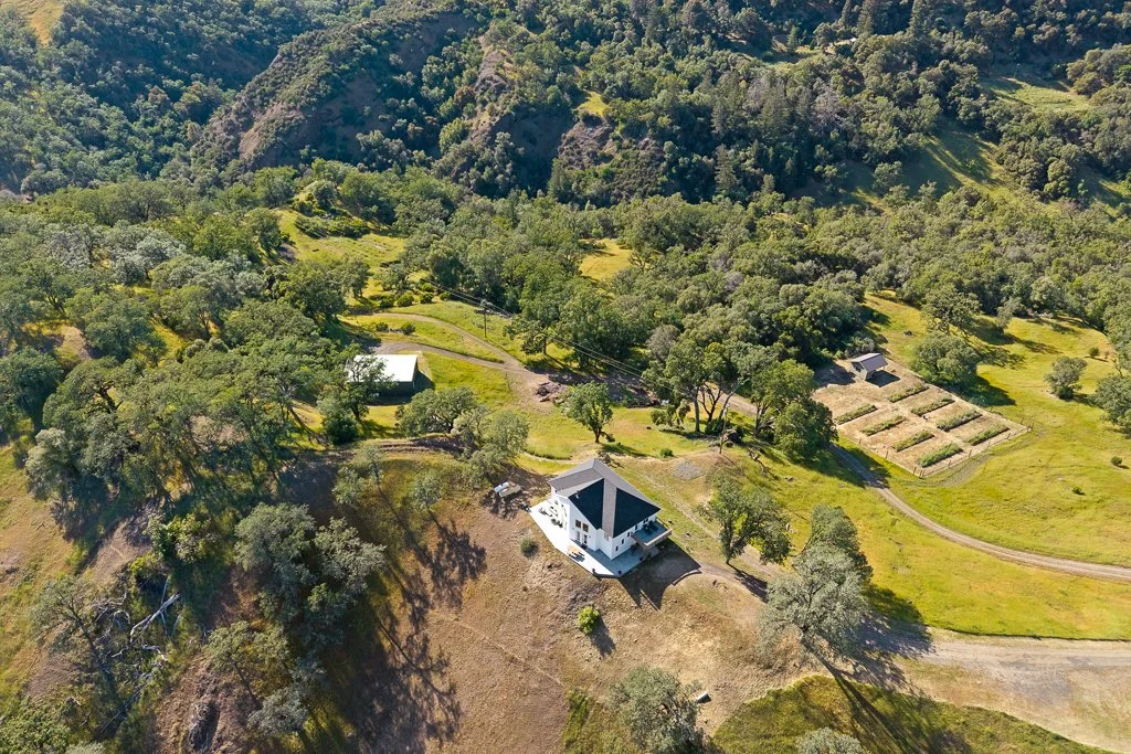 Aerial view of a rural landscape featuring green hills, trees, a white house with a dark roof, a few smaller outbuildings, and garden plots.