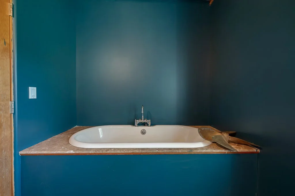A white bathtub with a faucet in front of a dark teal wall, placed on a marble countertop, with a trowel and a wooden board resting on the right side.