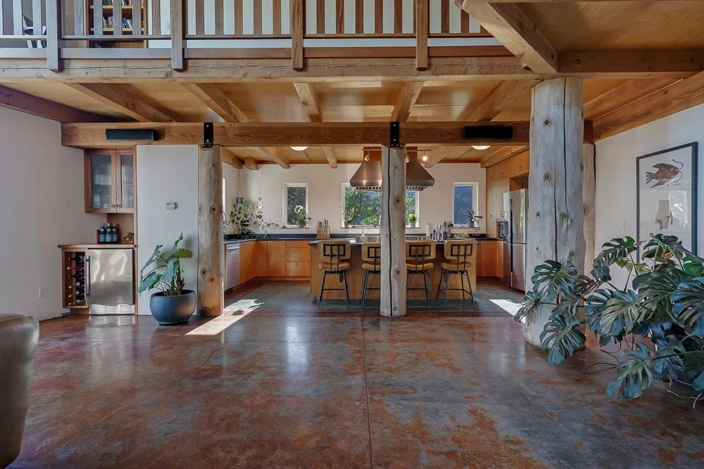 Open-concept kitchen with wooden cabinets, a center island with three bar stools, and large windows in a modern, rustic-style home. There's a staircase above, and indoor plants on both sides.