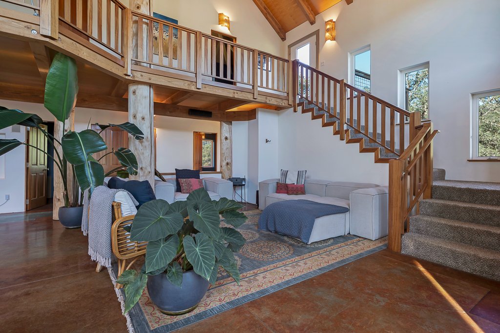 Living room with high vaulted ceilings, wooden beams, a staircase with wooden railing, large windows, and cozy furniture including a sectional sofa, armchair, and large potted plants.