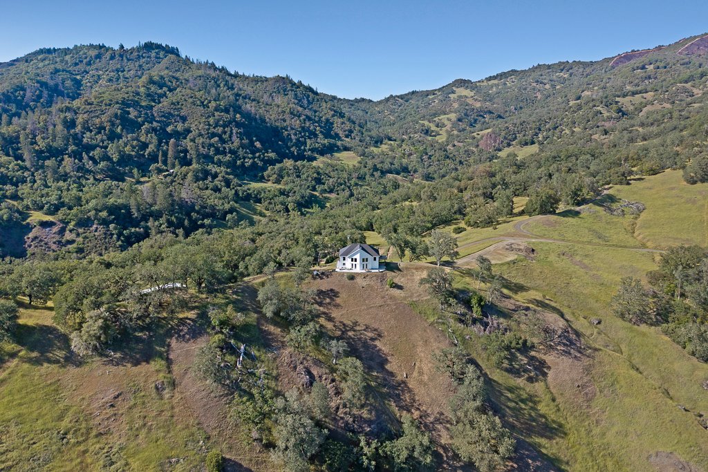 An aerial view of a landscape featuring a hillside with numerous trees and a single white house with a gray roof in the middle of the scene, set against a backdrop of tall, green mountains and a clear blue sky.
