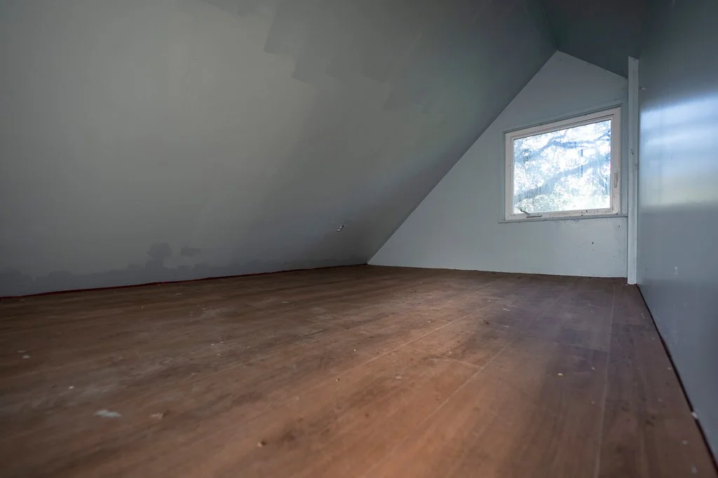Empty attic room with sloped ceiling, hardwood floor, and a single window showing trees outside.