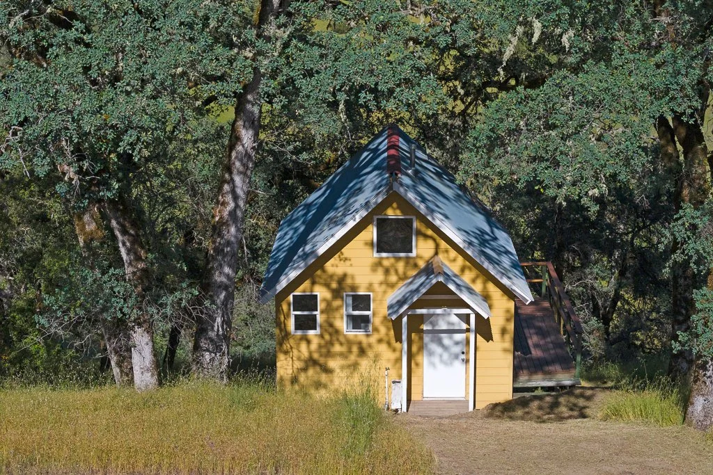 A small yellow house with a white door and several windows, surrounded by trees and grass, with a wooden deck on the right side.