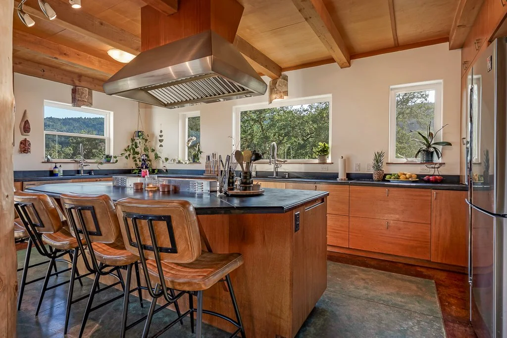 Modern kitchen with wooden cabinets, black countertops, a centralized kitchen island with bar stools, stainless steel range hood, and large windows showing a green outdoor landscape.