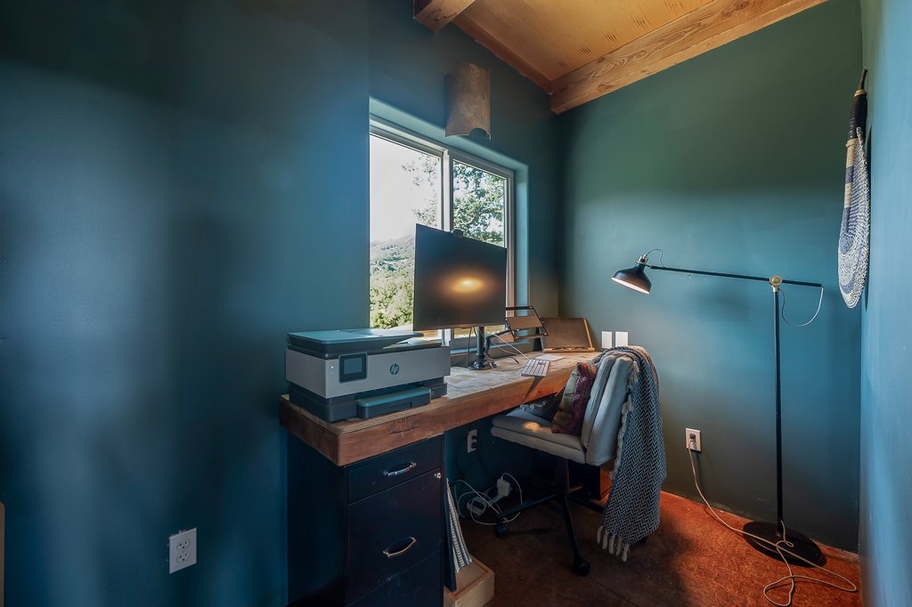 Home office with blue-green walls, wooden ceiling, window with outdoor view, black desk with printer, monitor, keyboard, and papers, gray chair with a pillow, black floor lamp, and hanging mitt to the right.