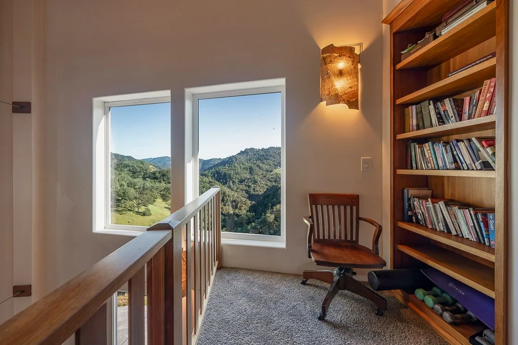 Home office corner with large windows overlooking a mountainous landscape, a wooden bookshelf with books and workout equipment, a wooden chair, and a wall-mounted lamp.