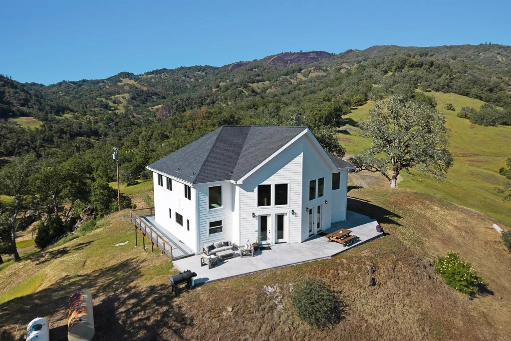 A white two-story house on a hillside with a large tree nearby, surrounded by green hills and mountains under a clear blue sky.