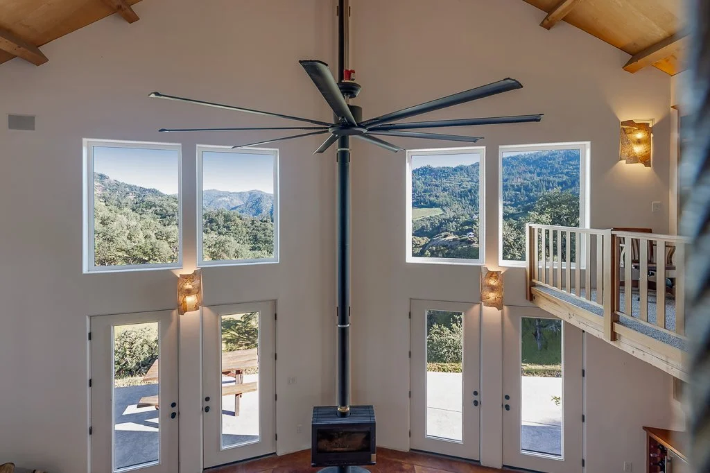 Interior view of a high-ceiling living room with large windows showing mountain views, a ceiling fan, and a balcony on the right.