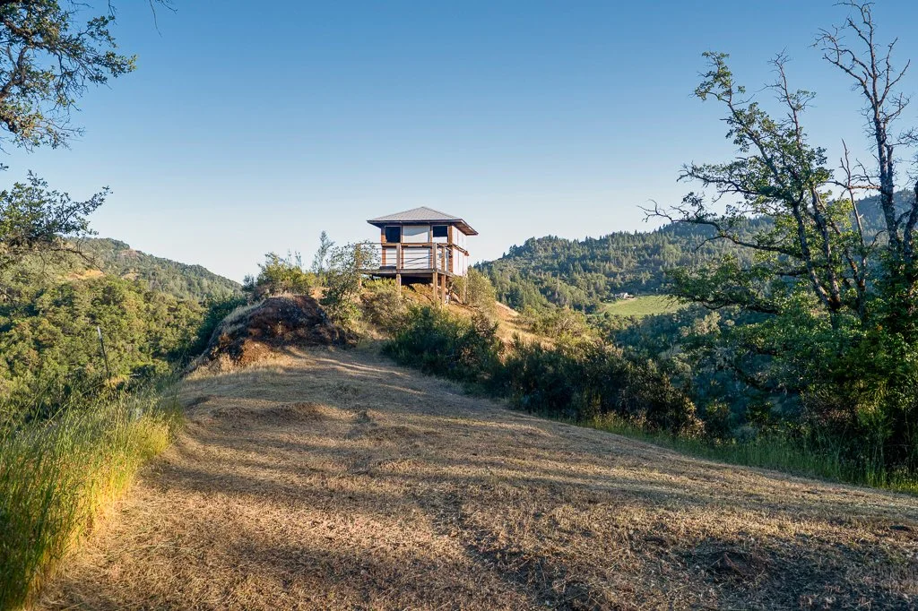 Wooden lookout tower on top of a dirt hill, surrounded by trees and rolling green hills under a clear blue sky.