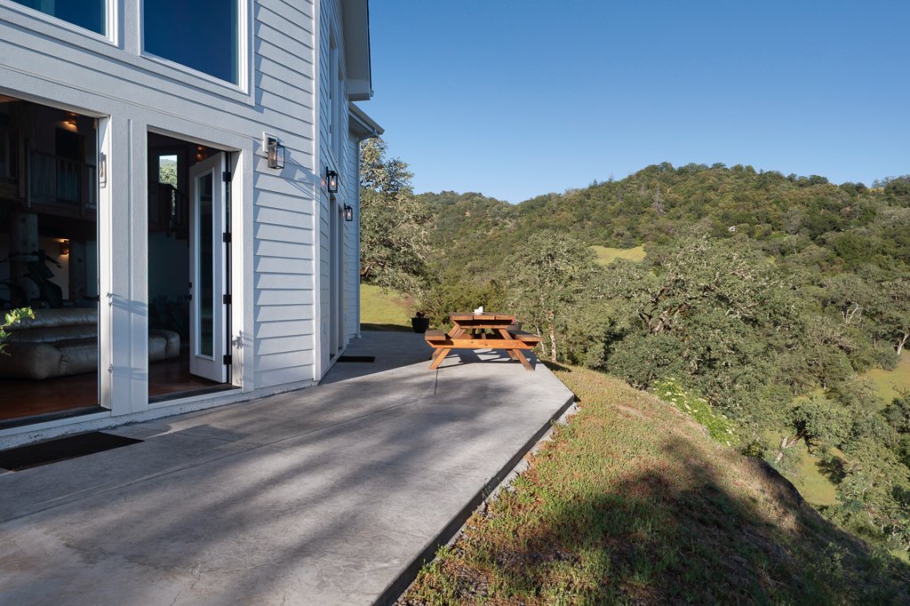 A house with white siding and glass doors opening to a concrete patio, with a wooden picnic table, overlooking a lush green hillside under a clear blue sky.
