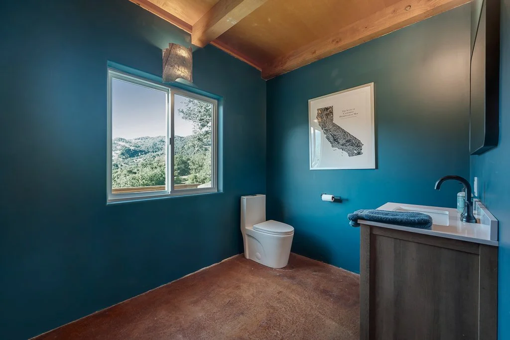 A bathroom with blue walls, a toilet, a window showing trees and mountains outside, a framed map of California on the wall, a small sink with a dark towel, and a ceiling with exposed wooden beams.