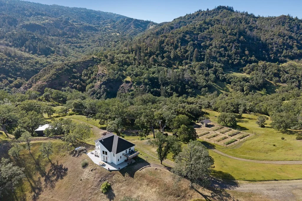 A white house with black roof shingles situated on a hillside surrounded by green trees and fields, with mountains in the background.