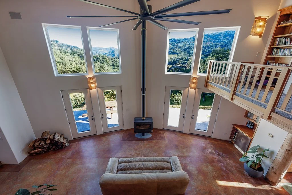 Interior view of a spacious living room with a high ceiling, large windows showing mountain scenery, a ceiling fan, a beige sofa, built-in bookshelves, and a wood stove, with sunlight streaming in.