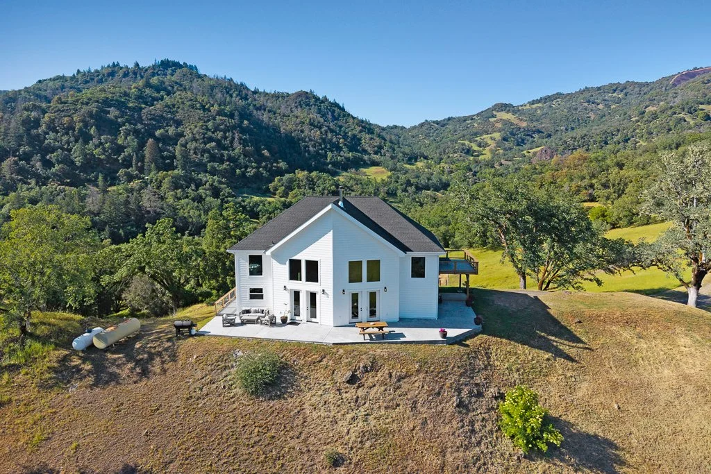 A white two-story house on a hillside overlooking mountains and trees, with a patio, outdoor seating, and propane tanks nearby.