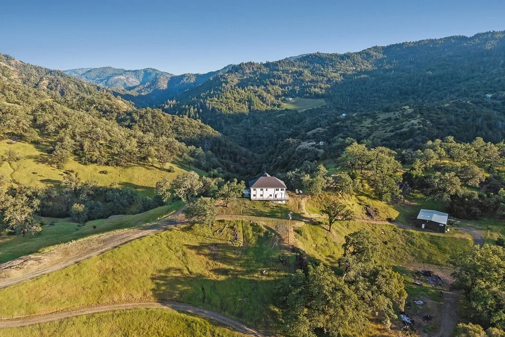 A house situated on a green hillside surrounded by trees, with mountains in the background and clear blue skies overhead.