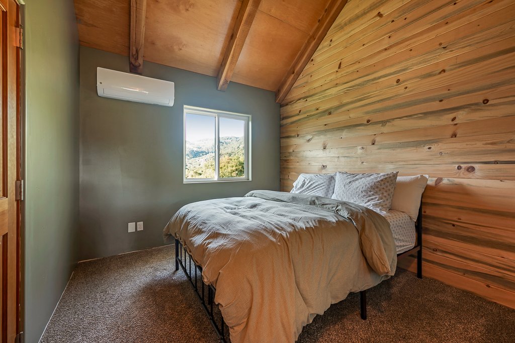 A bedroom with a sloped wooden ceiling, a window showing a mountain landscape, a bed with beige linens, and an air conditioning unit on the wall.