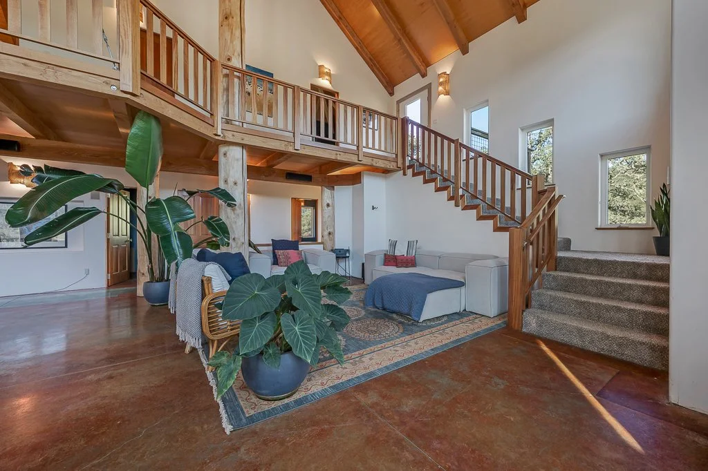 A spacious living room with a high wooden vaulted ceiling, large windows, a staircase, and a loft area. The room features a large potted plant, a white sectional sofa with pillows, and an ornate area rug.