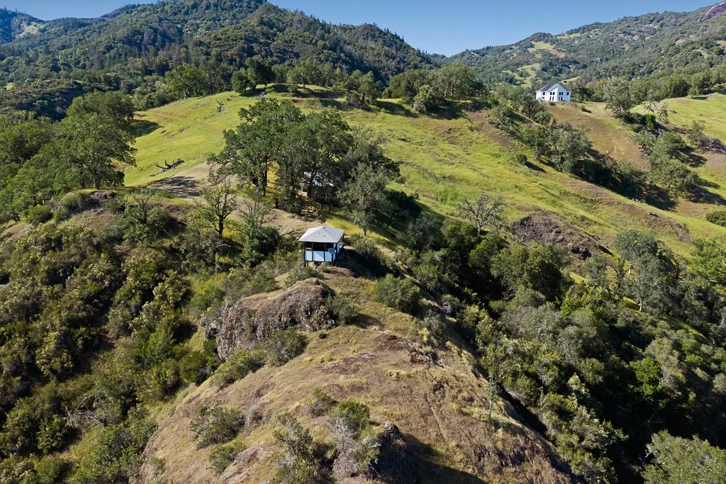 Aerial view of a hilly landscape with green trees and grass, featuring two houses on the slopes and distant mountains under a clear blue sky.