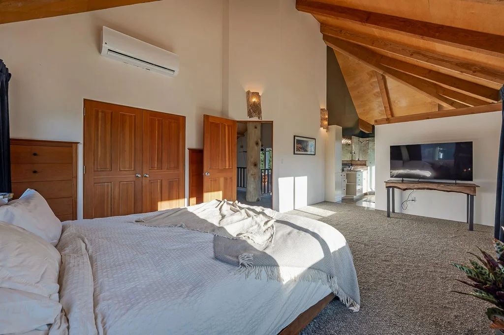 A bedroom with a large bed, wooden doors, a wall-mounted air conditioner, and a flat-screen TV on a stand. The room has vaulted wooden ceilings and a small open kitchen area in the background, with sunlight streaming in.