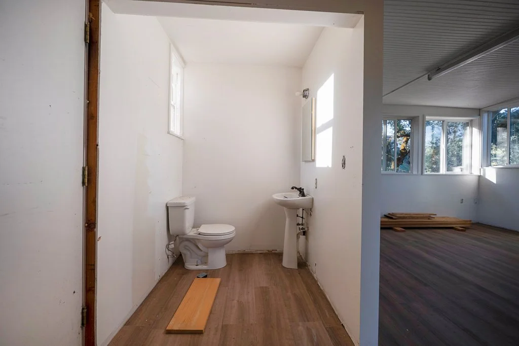 An unfinished small bathroom with a toilet and a pedestal sink, with a small window and a mirror, and a large room with windows and wooden flooring visible in the background.