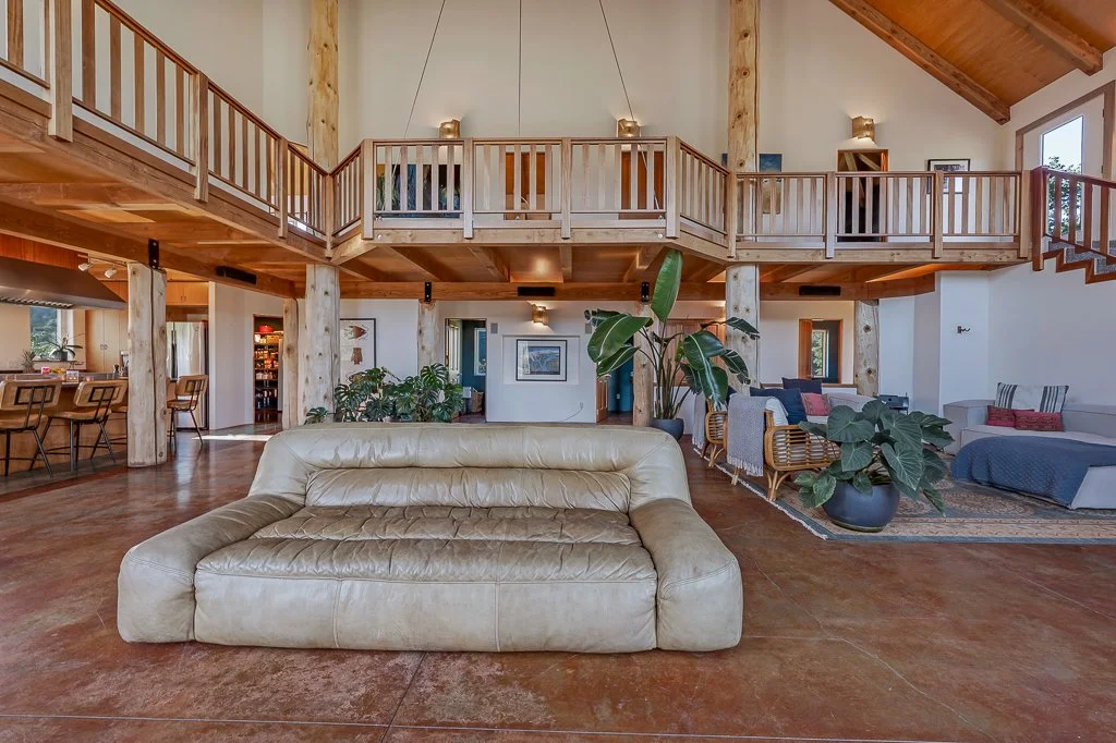 Spacious living room with a beige leather sofa in the foreground, large potted plants, and a cozy seating area on the right, with wooden beams and railing lining the second-floor balcony.