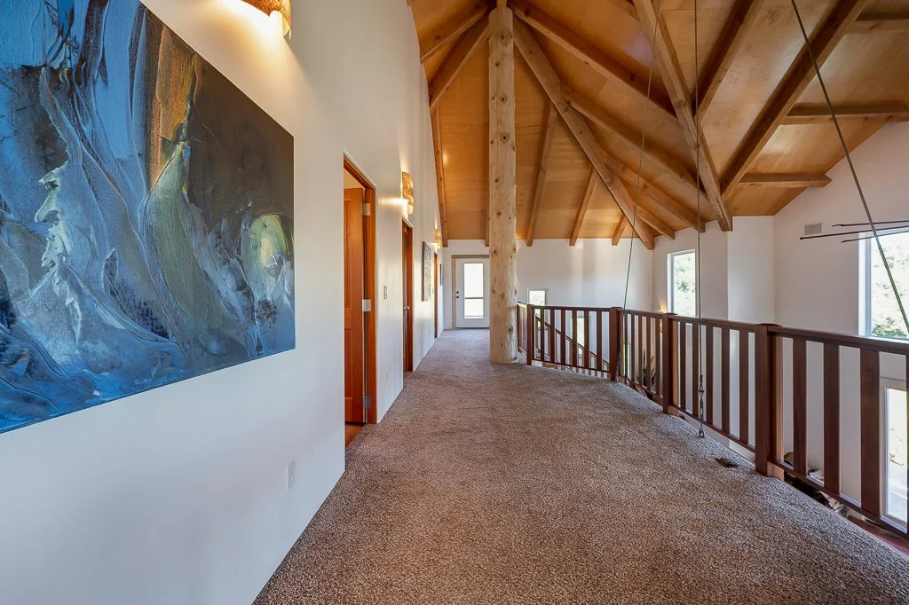 A hallway with a wooden ceiling and beams, featuring art on the wall, a wooden railing, and windows allowing natural light.
