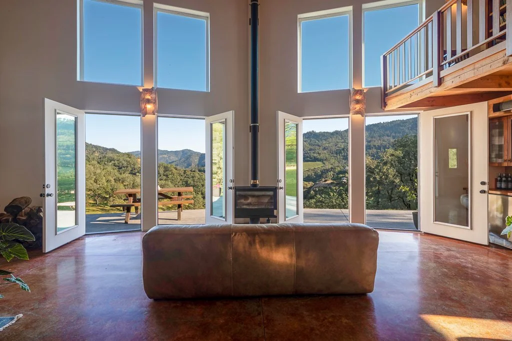 Living room with glass doors and large windows showing scenic mountain and greenery view, leather sofa, wood staircase, and balcony.