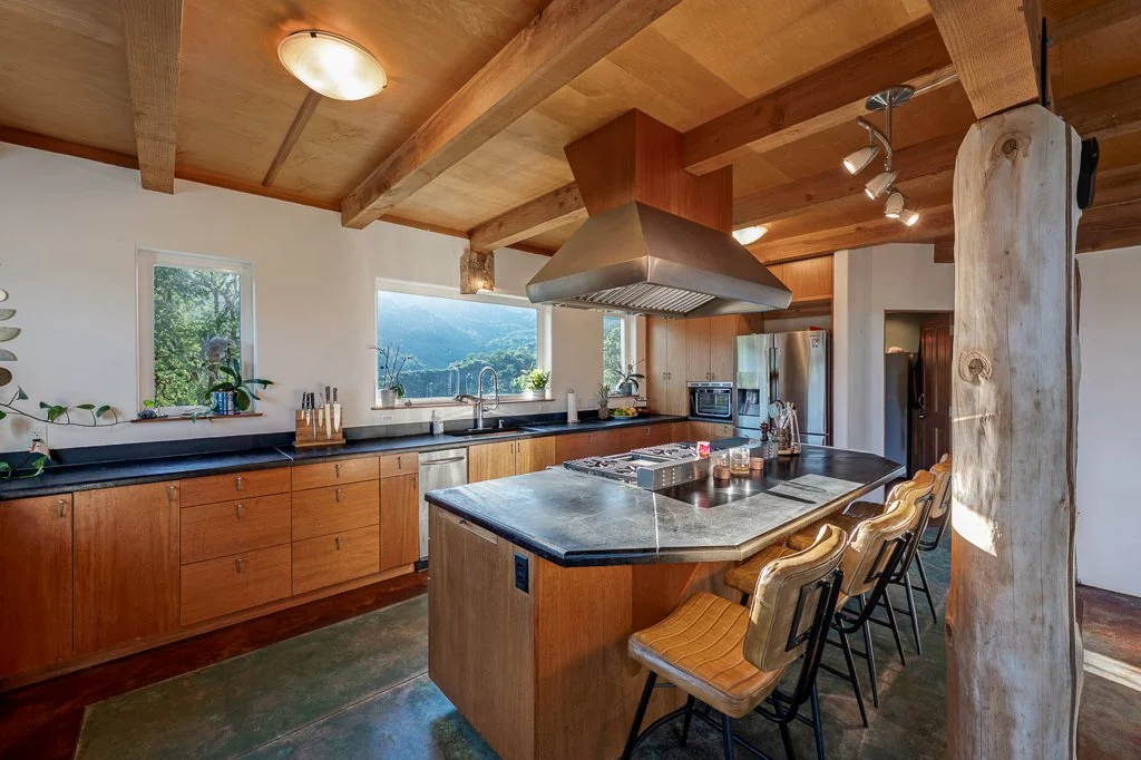 Kitchen with wood cabinets, stainless steel range hood, black countertops, and barstools around an island, featuring large windows with scenic mountain views.