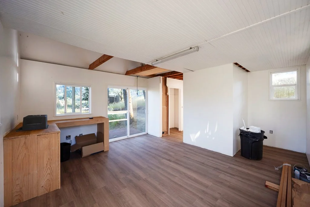 Empty room with white walls and wooden floors, several windows, partial ceiling and wall damage, and some furniture including a black trash can.