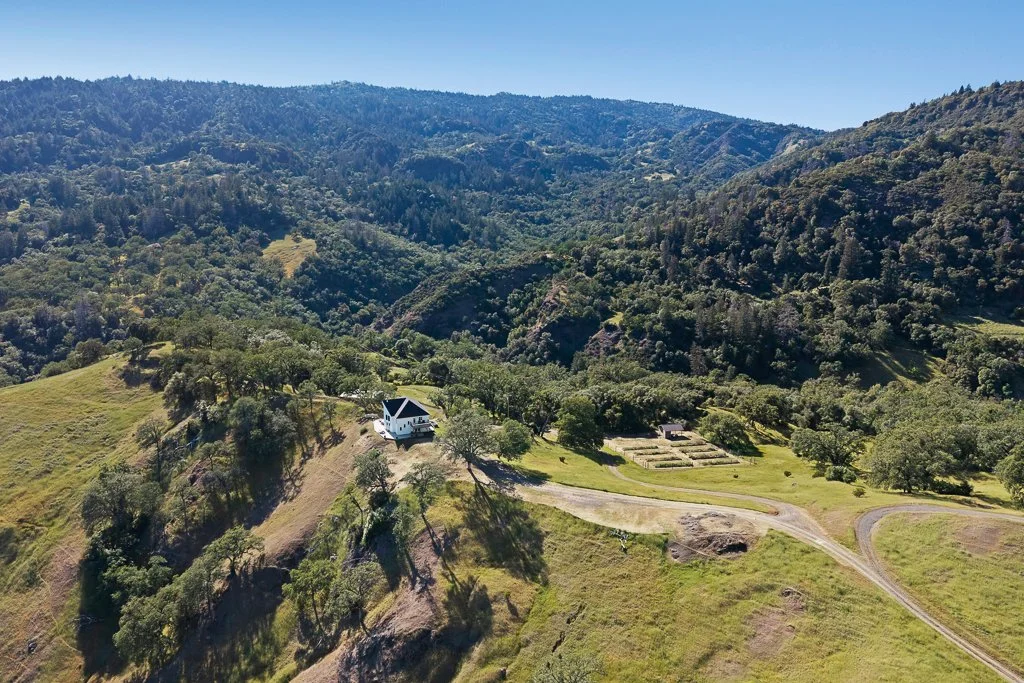A white house with a black roof sits on a grassy hillside surrounded by trees, with a winding dirt road leading to it, nestled in a valley of rolling green hills and forested mountains in the background.